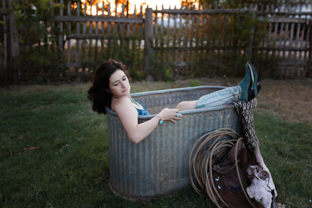 Thalia lying back in a tub, with cowgirl boots and rustic props around, under the Texas sunset.